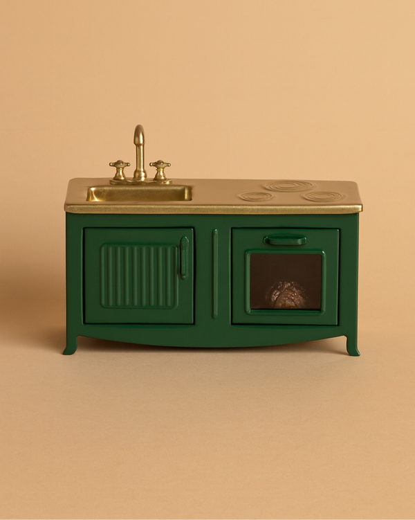 The Maileg Kitchen - Dark Green is a miniature toy oven with a gold faucet, sink, and stovetop, plus an oven holding a small loaf of bread. Ideal for a mouse family's metal kitchen. Set against a solid beige background.