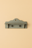 The Maileg Miniature Dollhouse Shelf is a small, decorative wooden shelf with two knobs and a scalloped top edge, painted in muted blue-green—perfect for Maileg dollhouse decor. Displayed against a beige background.