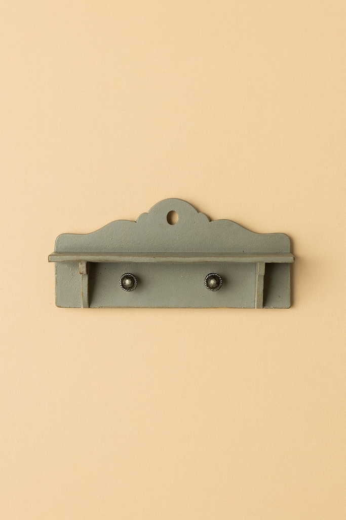 The Maileg Miniature Dollhouse Shelf is a small, decorative wooden shelf with two knobs and a scalloped top edge, painted in muted blue-green—perfect for Maileg dollhouse decor. Displayed against a beige background.