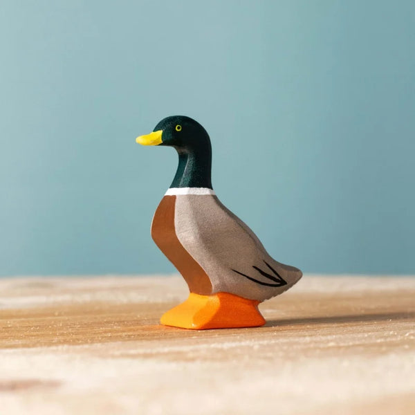 The Bumbu Handmade Wooden Mallard Duck, featuring a green head, yellow beak, and orange feet, stands on a light wooden surface with a plain blue background.