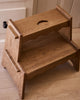 The Häti at Home - Margot Step Stool, a compact solid oak stool with two steps and a handle cutout on the top step, sits on a light wood floor next to a cabinet drawer.
