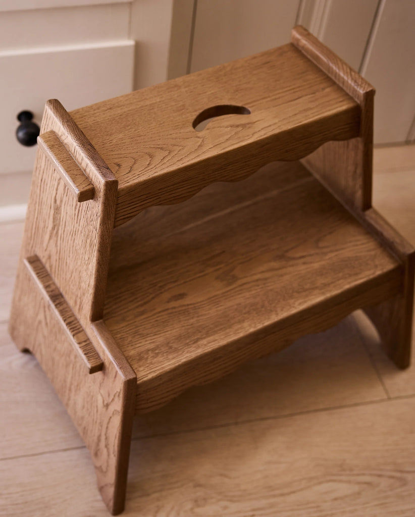 The Häti at Home - Margot Step Stool, a compact solid oak stool with two steps and a handle cutout on the top step, sits on a light wood floor next to a cabinet drawer.