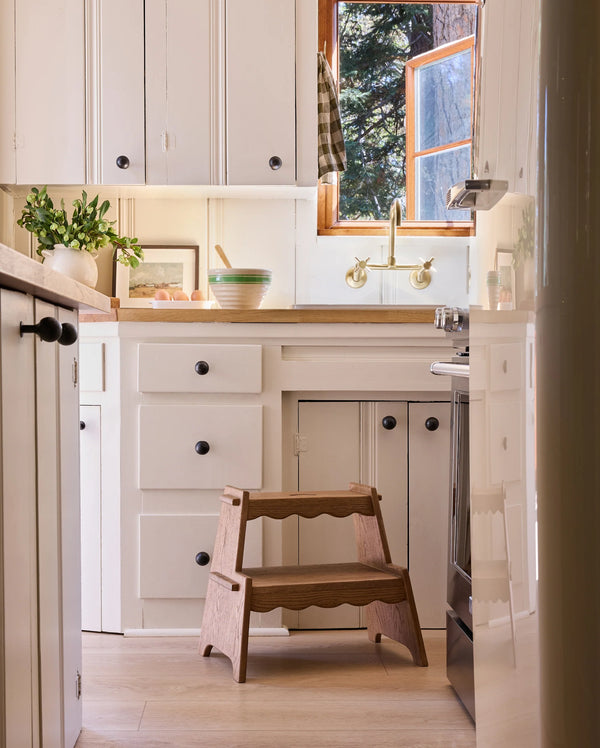 A cozy kitchen with white cabinets, wooden countertops, and a window above the sink. The Häti at Home Margot Step Stool sits on the light wood floor, while greenery and kitchenware add charming, homey touches.