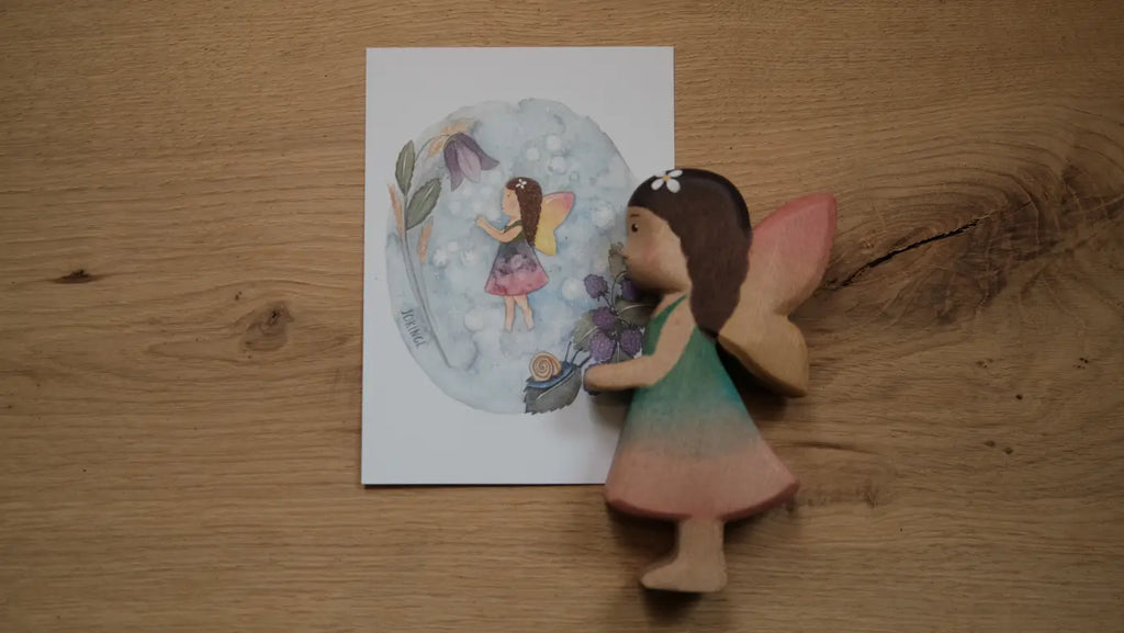 The Handmade Holzwald Maya Fairy and Postcard features a handcrafted wooden fairy figurine resting on a wooden surface beside a card adorned with an illustration of the same fairy, complete with wings, dress, and brown hair surrounded by flowers—making it a charming addition to any natural wooden toy collection.