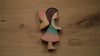 A Handmade Holzwald Maya Fairy and Postcard with brown hair, pastel wings, and a dress in shades of blue, green, and pink rests on a light wooden surface. This enchanting piece highlights the artistry of handmade wooden figures and brings a magical touch to any collection.
