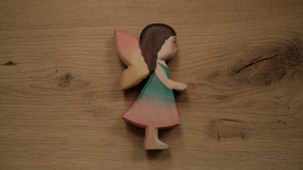 A Handmade Holzwald Maya Fairy and Postcard with brown hair, pastel wings, and a dress in shades of blue, green, and pink rests on a light wooden surface. This enchanting piece highlights the artistry of handmade wooden figures and brings a magical touch to any collection.