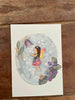 The Handmade Holzwald Maya Fairy and Postcard features a watercolor illustration of a fairy with butterfly wings standing among purple flowers, leaves, and a snail—all set against a circular blue background on a wooden surface—capturing the enchanting essence of natural wooden toys and handcrafted figures.