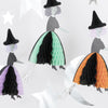 The image showcases the Meri Meri Flying Honeycomb Witch Decorations, featuring three whimsical witches with colorful, pleated skirts in green, orange, and purple. Each witch is holding a broomstick, wears a black hat, and has an illustrated face. They float among white stars and moons—perfect for adding a festive touch to your Halloween party decor. Note that this item is on final sale.