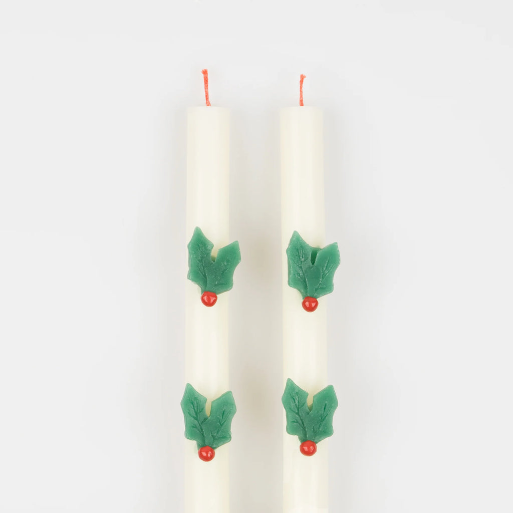 The MeriMeri Holly Table Candles, a set of two white taper candles with red wicks, are beautifully decorated with green holly leaves and red berries. These classic Christmas candles stand upright against a simple white background, capturing the essence of traditional decorations that add festive warmth to any space.