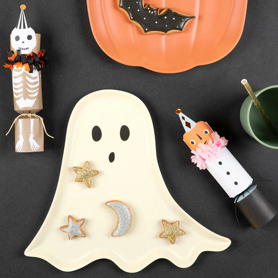 A Halloween-themed setup featuring a Meri Meri Reusable Bamboo Ghost Plate with star and moon cookies, a pumpkin-shaped bamboo plate, two skeleton and clown-themed party favors, and a black cup with a green and white striped straw, all perfect for Halloween parties on a black surface.