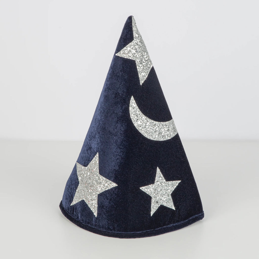 The Meri Meri Blue Velvet Wizard Costume - Final Sale features a cone-shaped velvet hat in dark blue, adorned with silver glitter stars and a crescent moon, standing upright on a plain light surface. Perfect for magical dress-up!.