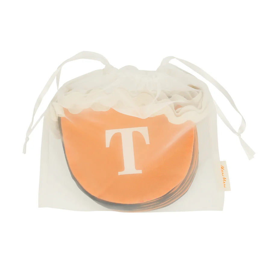 A semi-transparent drawstring bag holding a stack of circular orange coasters, each featuring a large white letter "T" on the top coaster, adds an air of mystery perfect for any Halloween setup. The white drawstring closure and small tag with text enhance the intrigue of the Meri Meri Trick Or Treat Fabric Garland.