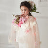 A young girl with dark hair stands against a white background, thoughtfully holding pink flowers while wearing the Meri Meri Flower Cape—an embellished kids cape perfect for floral dress-up. Final Sale.