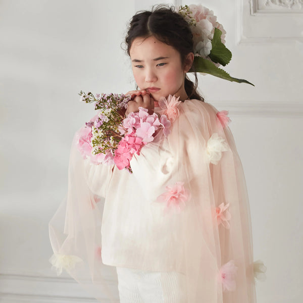 A young girl with dark hair stands against a white background, thoughtfully holding pink flowers while wearing the Meri Meri Flower Cape—an embellished kids cape perfect for floral dress-up. Final Sale.