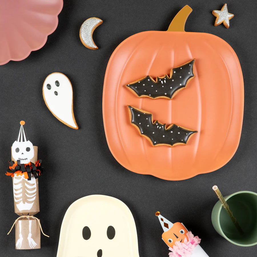 A Halloween-themed flat lay featuring pumpkin-shaped and ghost-shaped plates, bat and ghost cookies, a paper skeleton decoration, a green cup with a pink cupcake, a crescent moon, a star, and festive party items on a black background. Highlighting the display is the Meri Meri Reusable Bamboo Pumpkin Plate, perfect for spooky gatherings.