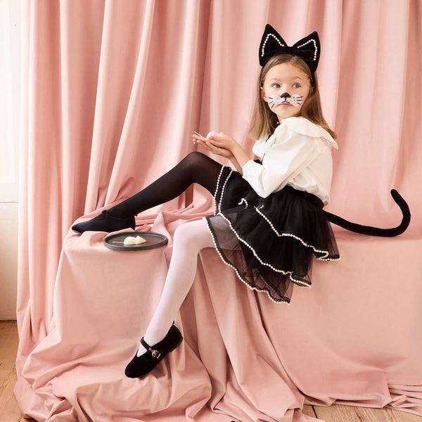 A young girl wears the Meri Meri Cat Costume—complete with cat ears headband, face paint, and black tulle skirt—while sitting on a pink draped surface and posing playfully like a cat, a plate beside her.