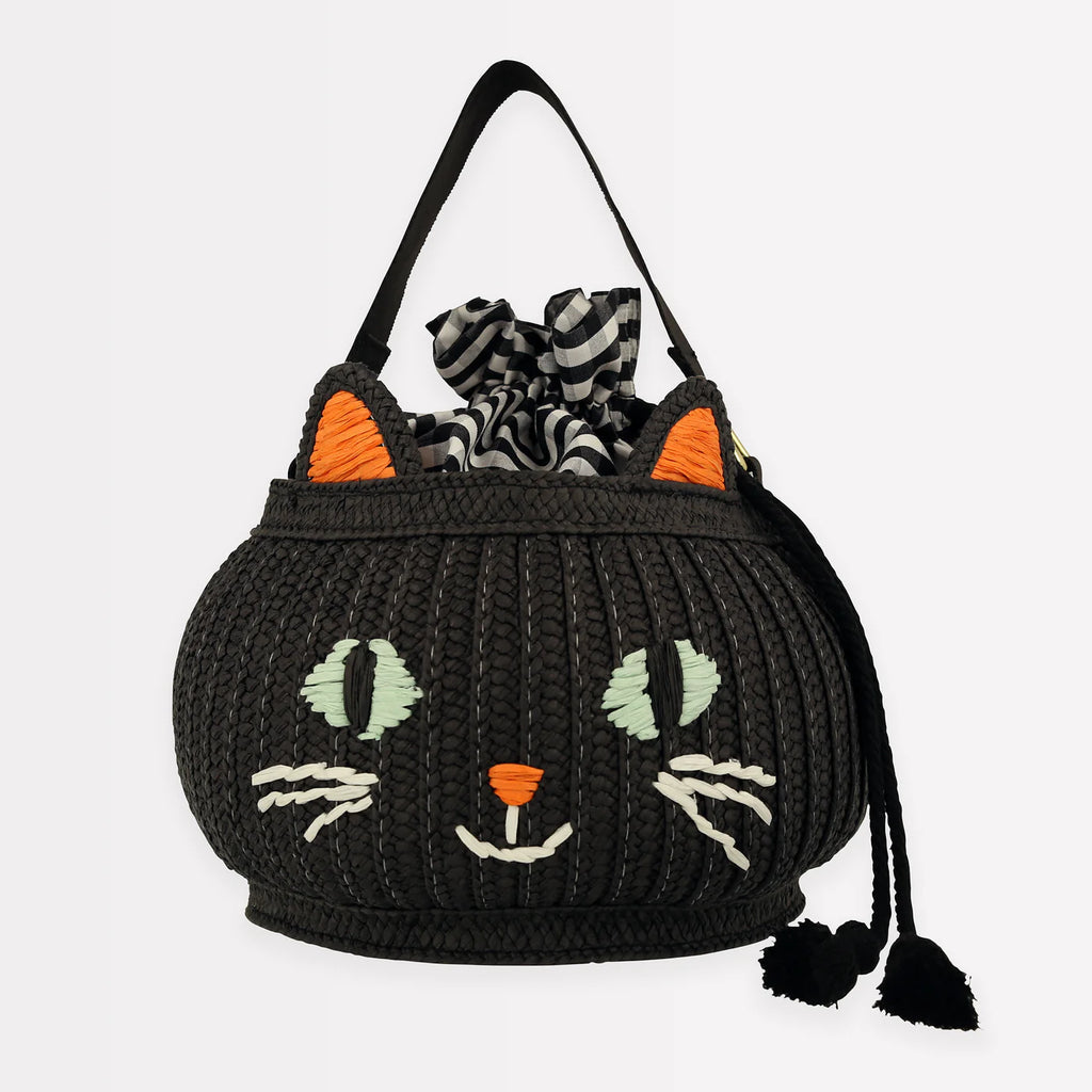 The Trick Or Treat Cat Basket is a black woven handbag designed to resemble a cat's face, featuring large green eyes, white whiskers, and orange ears. Perfect for collecting treats, it has black shoulder straps and is accented with a black and white checkered cloth emerging from the top.