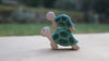 Two Handmade Holzwald Sea Turtles with painted green shells are stacked on a light surface, with a blurred grassy and tree-filled outdoor background.