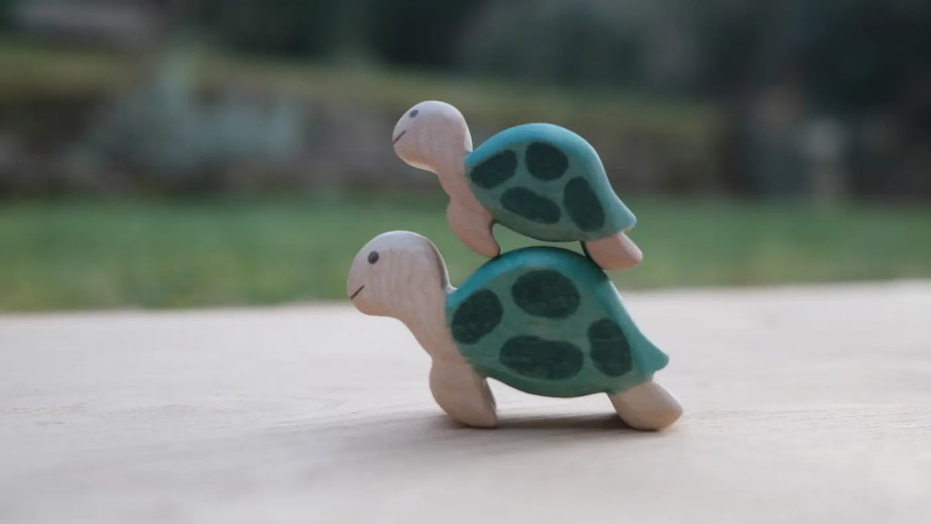Two Handmade Holzwald Sea Turtles with painted green shells are stacked on a light surface, with a blurred grassy and tree-filled outdoor background.