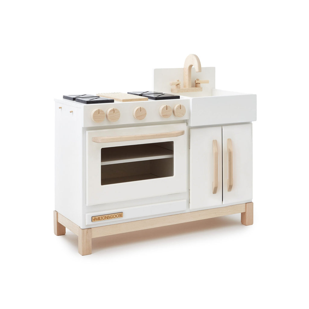 The Milton & Goose Essential Play Kitchen, made in the USA, features a white and natural wood finish with a stove, knobs, oven, sink with faucet, and cabinet doors—perfect for imaginative play.