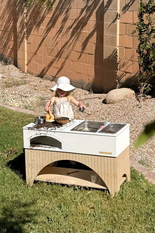 A young child in a white sunhat and light-colored dress is playing with a Milton & Goose Mud Kitchen (Made in USA) in the backyard. The outdoor playset includes a sink and stove with pots and toy food. The child is focused on the play, while the sun casts shadows on a brick wall behind.