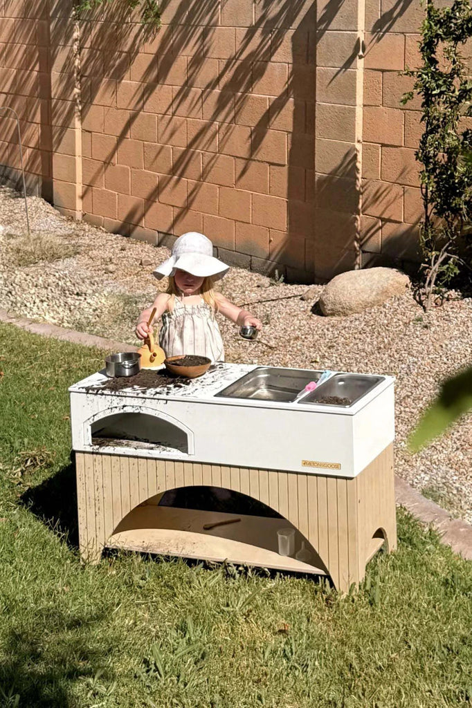 A young child in a white sunhat and light-colored dress is playing with a Milton & Goose Mud Kitchen (Made in USA) in the backyard. The outdoor playset includes a sink and stove with pots and toy food. The child is focused on the play, while the sun casts shadows on a brick wall behind.