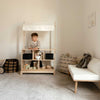 A young child stands behind the Milton & Goose Market Stand in a cozy, neutral-toned room with a small white sofa, plush bear, and baskets—a bright, minimalistic setup ideal for imaginative kids’ play shop fun.