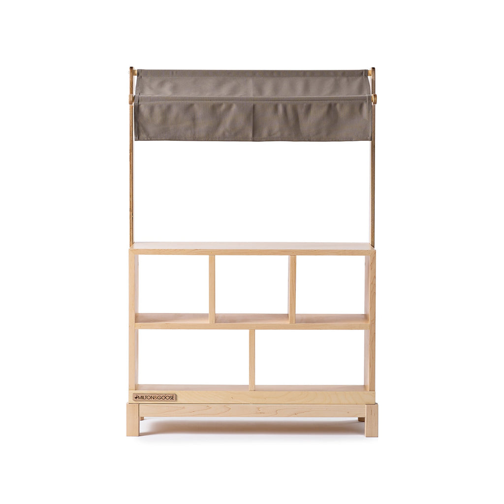 The Milton & Goose Market Stand features a handcrafted wooden frame with a light wood finish, an open shelf design, four compartments, and a gray fabric canopy—ideal for any kids' play shop with its simple, modern look.