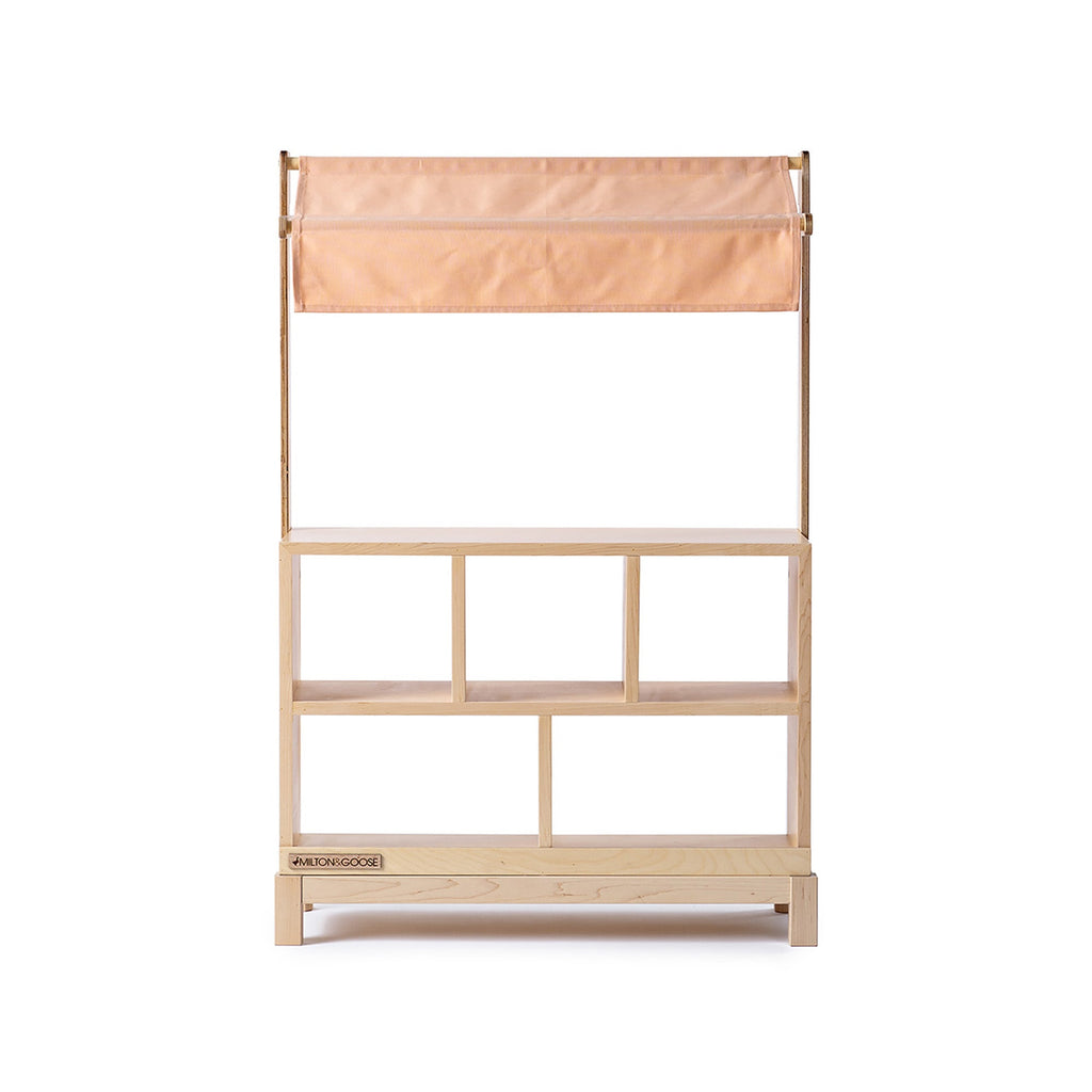 The Milton & Goose Market Stand features open shelves and a light peach fabric canopy, making it a perfect kids’ play shop. Handcrafted and photographed against a white background.
