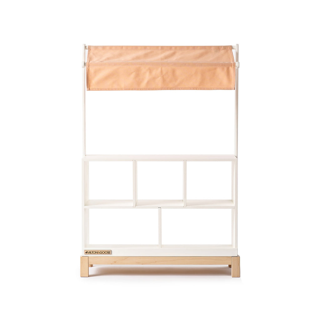 The Milton & Goose Market Stand features a white and light wood design with four open shelves and a light tan fabric canopy, displayed against a plain white background.
