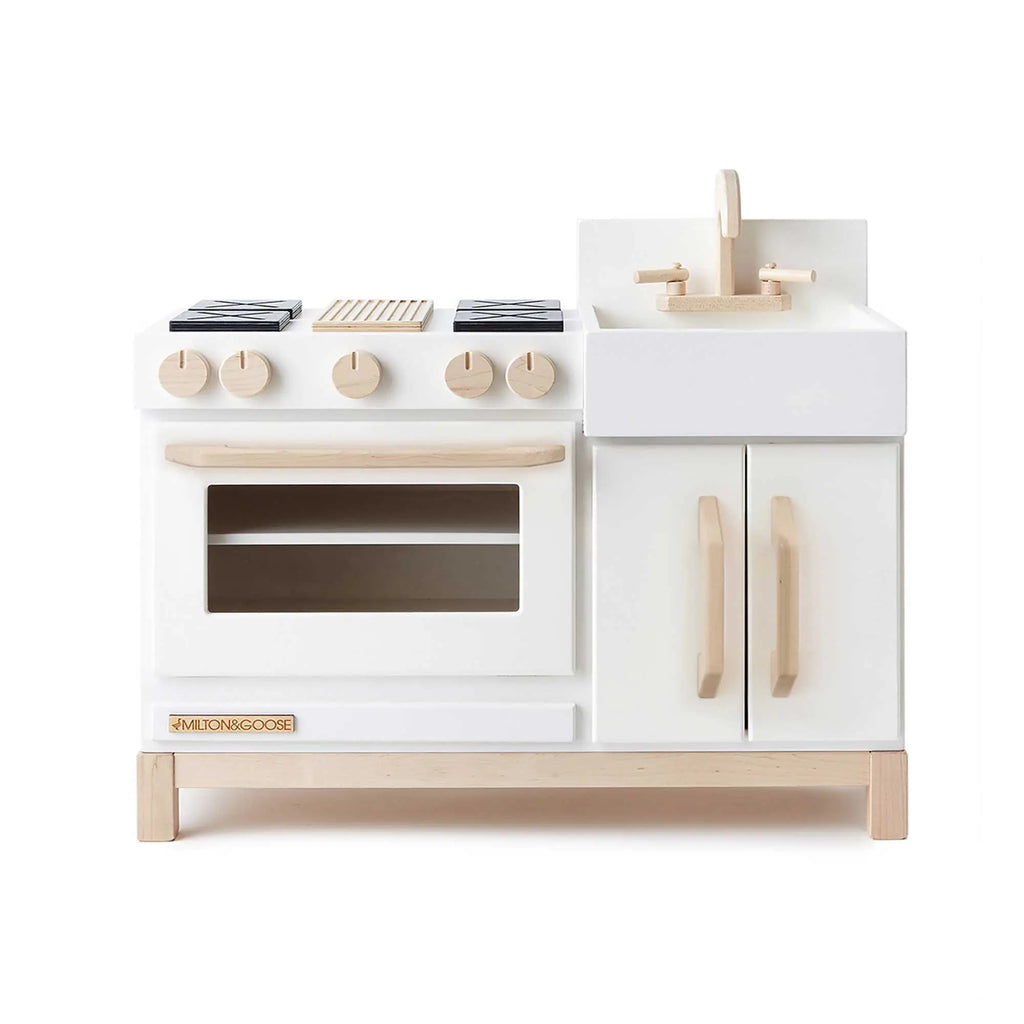 The Milton & Goose Essential Play Kitchen, handcrafted in the USA with white and light wood, features four burners, a clear-door oven, sink with faucet, and cabinets—ideal for imaginative play.