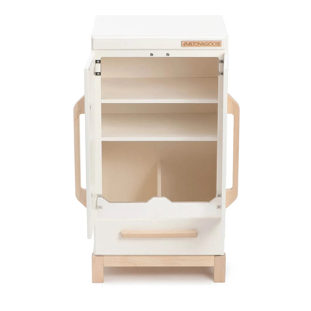 The Milton & Goose Essential Play Refrigerator—made in the USA—features a white finish, wooden handles, an open door showing two shelves above a large compartment, and stands on a wooden base, ideal for play food or any imaginative kitchen setup.