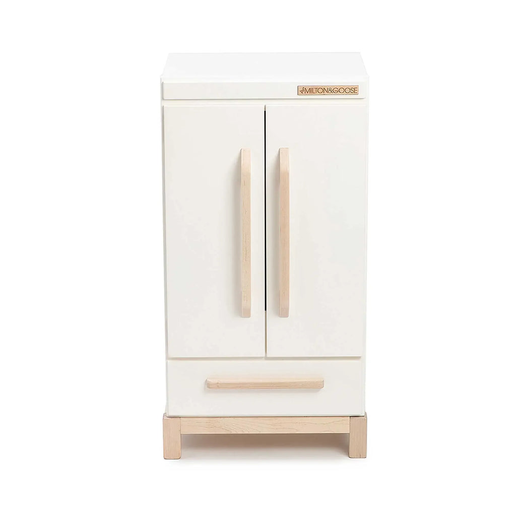 The Milton & Goose Essential Play Refrigerator—Made in the USA—is a small, white wooden cabinet with two vertical handles and a bottom drawer on light wooden legs, perfect for a play kitchen and hours of imaginative play.