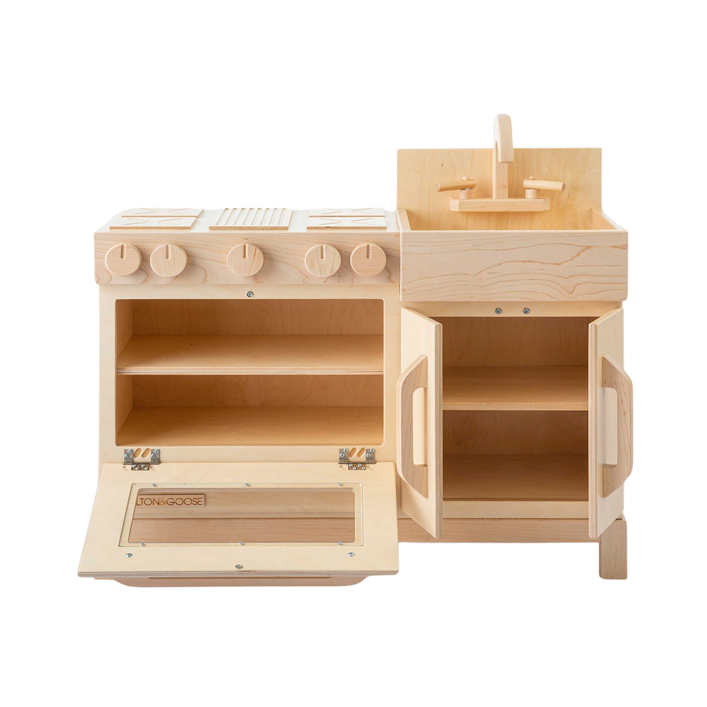 The Milton & Goose Essential Play Kitchen, made in the USA from light-colored wood, features a stovetop with knobs, an oven with opening door, shelves, and a sink with faucet—perfect for imaginative play.
