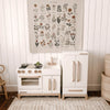 A Milton & Goose Essential Play Refrigerator, made in the USA, stands against a white paneled wall with a stove, sink, play food, and a botanical poster above. A woven basket sits on the right—perfect for imaginative play.