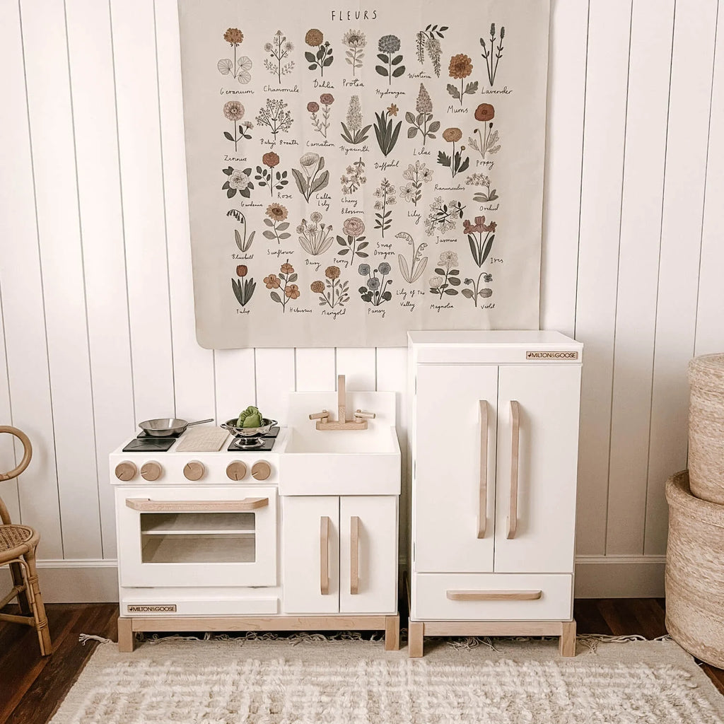 A Milton & Goose Essential Play Refrigerator, made in the USA, stands against a white paneled wall with a stove, sink, play food, and a botanical poster above. A woven basket sits on the right—perfect for imaginative play.