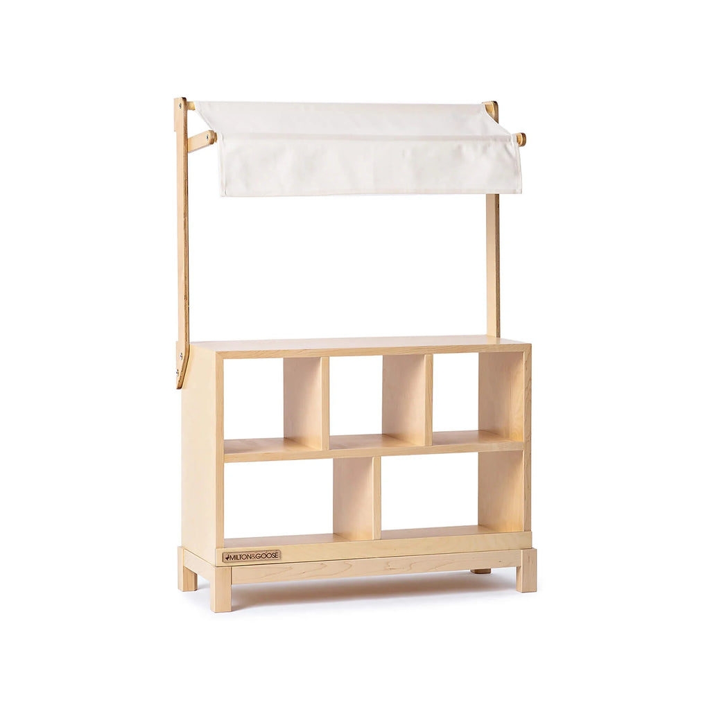 The Milton & Goose Market Stand is a handcrafted play stand featuring six open cubby shelves and a white fabric canopy, perfect for children’s play or storage, shown against a white background.