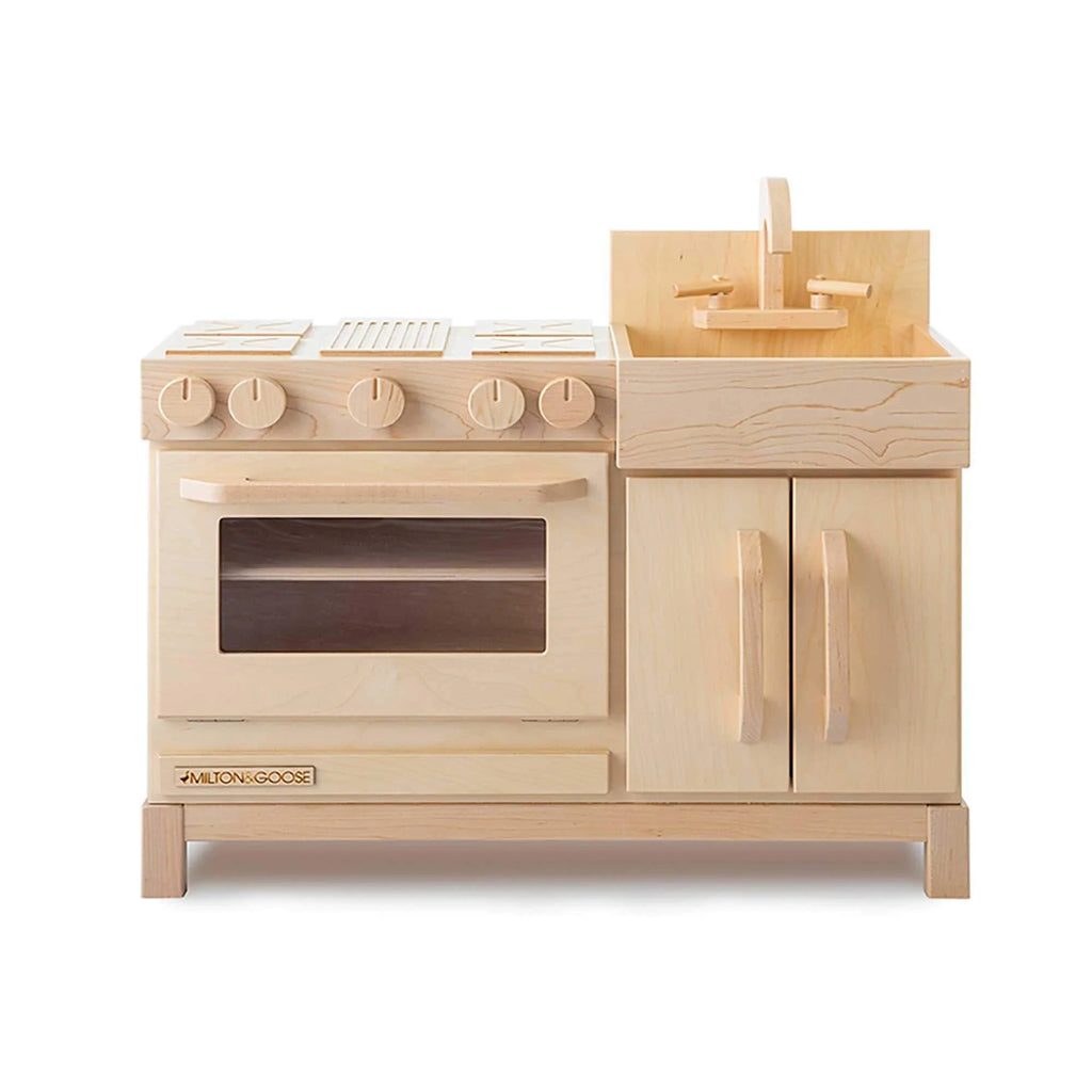 The Milton & Goose Essential Play Kitchen—made in the USA—features an oven with control knobs, stove, sink with faucet, and double-door cabinets crafted from light-colored wood to inspire imaginative play.