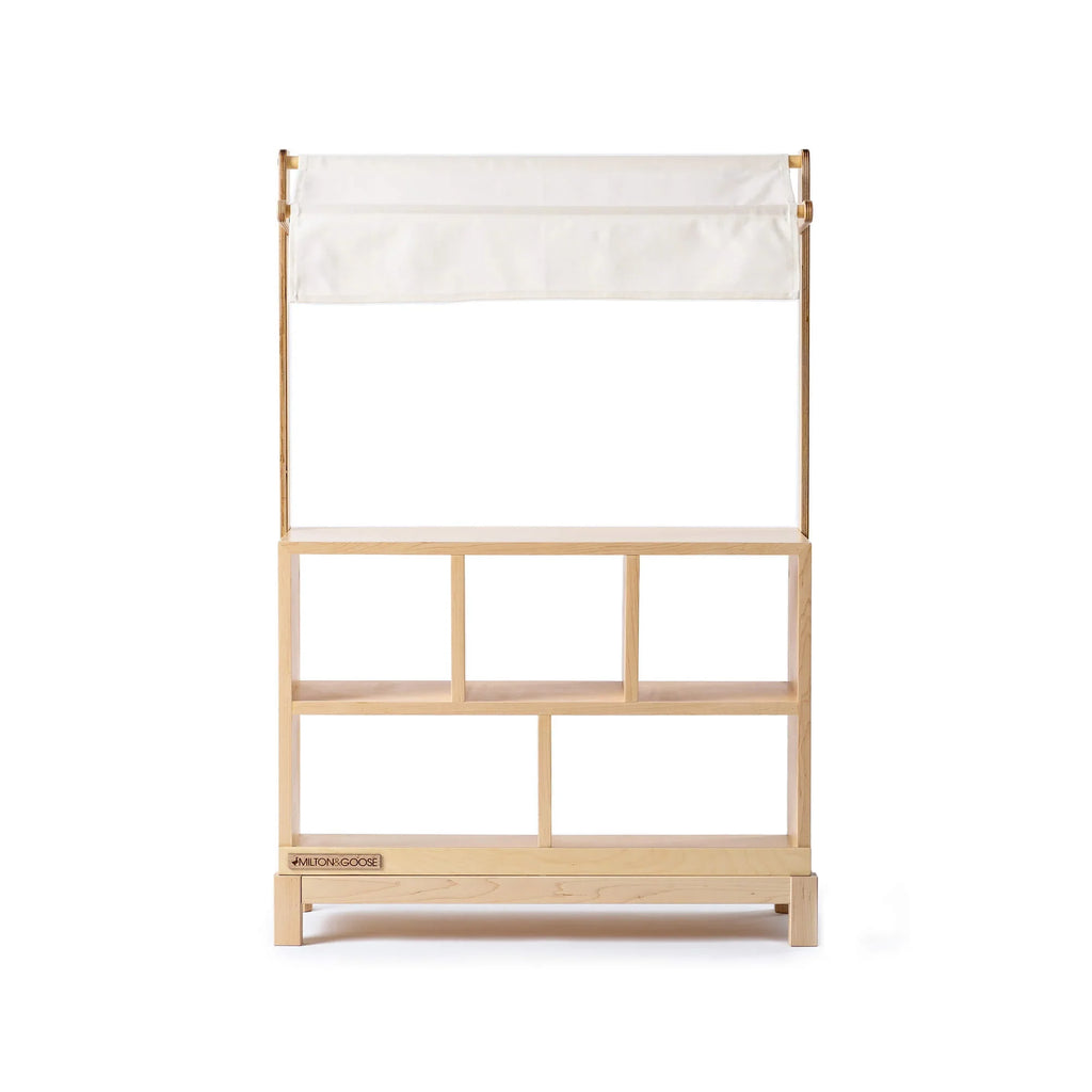 The Milton & Goose Market Stand features a white fabric canopy, open shelves, and a platform base, designed to inspire imaginative play—perfect for use as a kids' play shop.