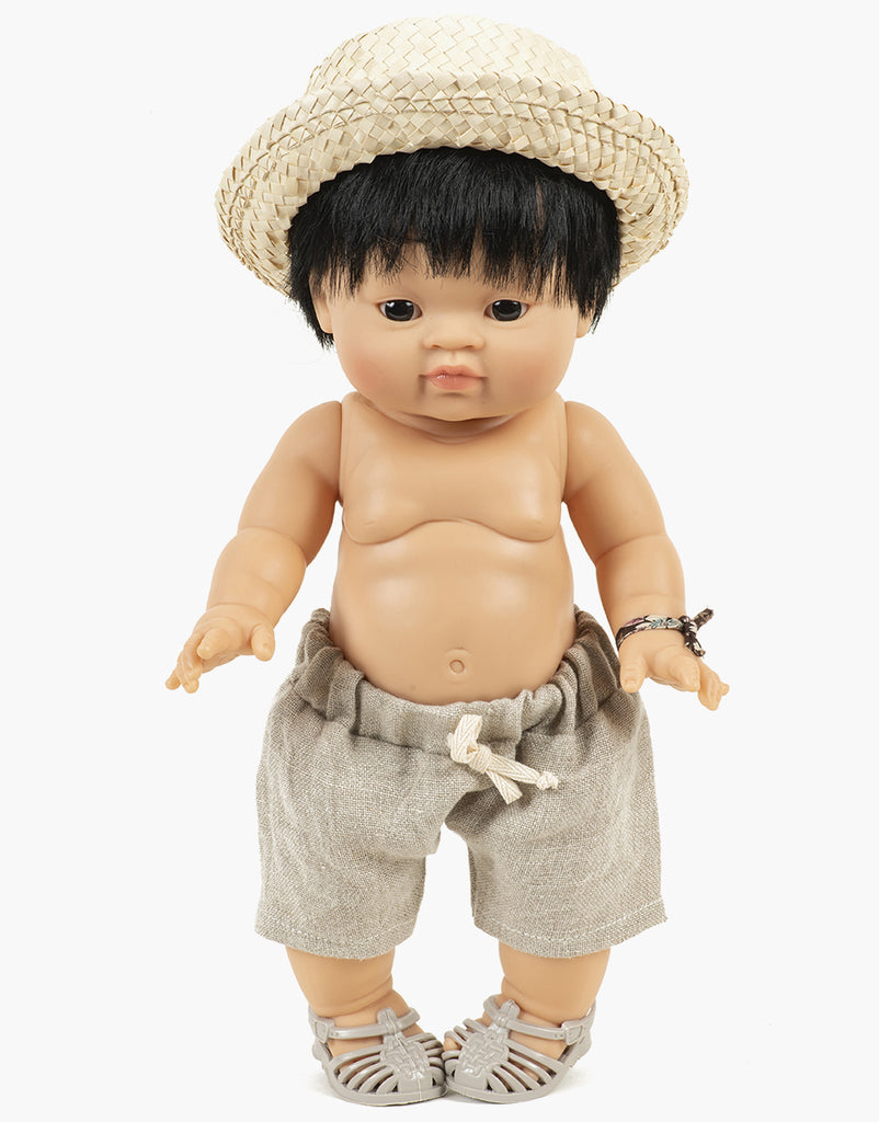 A Minikane | Doll Straw Hat with a light tan complexion and short black hair wears a Pablo straw hat, beige shorts with a drawstring, and gray sandals. Designed in France, the doll's arms are slightly extended, and it wears a small bracelet on its right wrist.