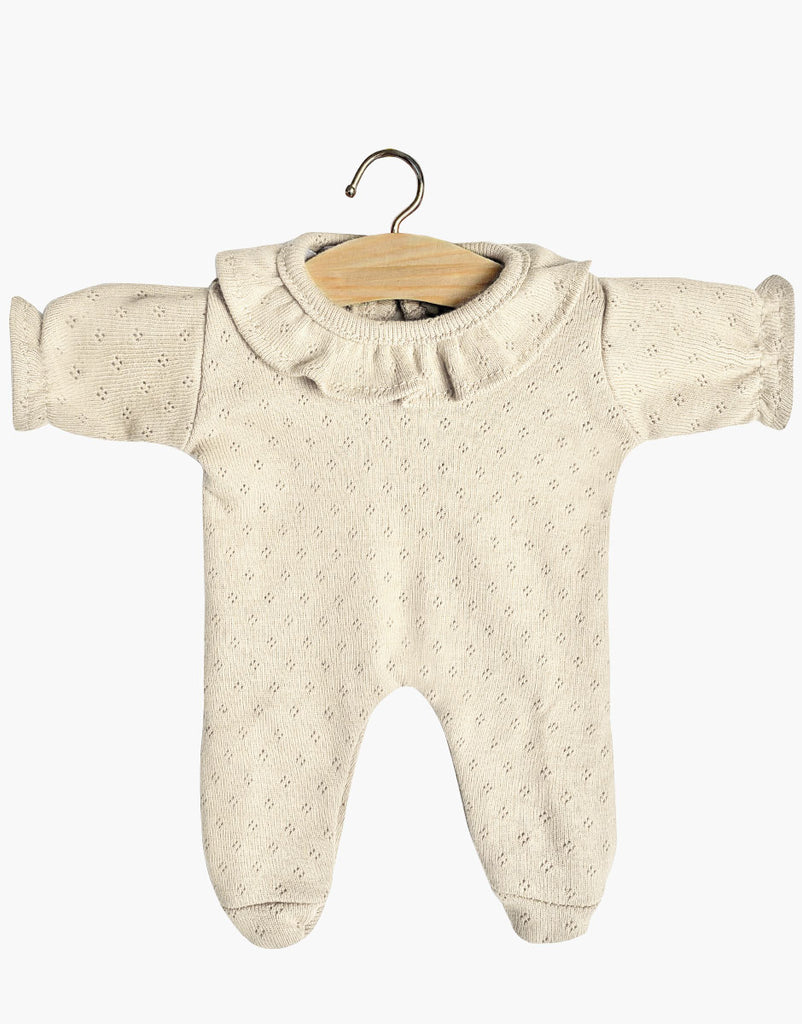 A cream-colored Minikane Doll Clothing | Sleep Well Dotted Linen for Minikane Babies clothes hangs on a wooden hanger. The onesie has long sleeves, footed legs, and a ruffled collar with delicate eyelet detailing throughout the fabric. Perfect for 11" dolls, this charming piece is made in France.