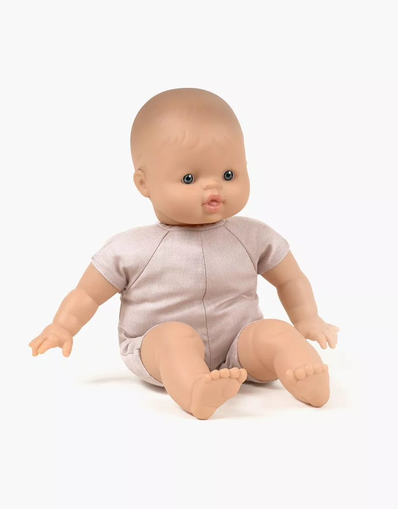 The Minikane Soft Body Doll (11") - Madder features a smooth bald head, blue eyes, and slightly open lips. The doll is dressed in a light pink outfit and sits upright with legs extended forward and arms resting at its sides. Made from phthalate-free vinyl, it's perfect for imaginative playtime against a plain white background.