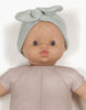 A doll with a smooth light brown complexion is dressed in a light pink outfit from Collégien and wears the Minikane Doll Clothing | Green Tea Doll Headband. The Minikane doll has wide, expressive eyes and slightly open lips. The background is plain white.