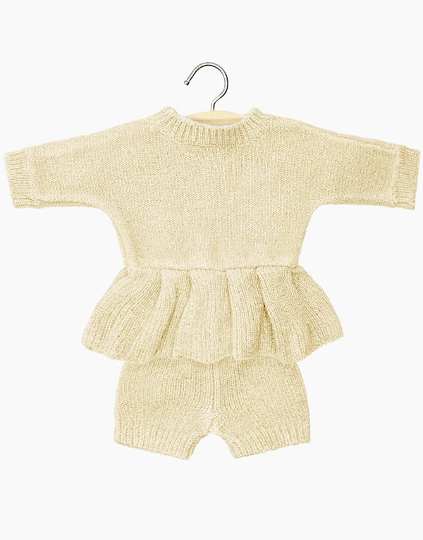 A knitted baby outfit in cream hangs on a hanger, similar to the Minikane Doll Clothing's Félicie Set. It includes a long-sleeve top with ruffled peplum and matching shorts made of cozy fabric, beautifully displayed on a plain white background.