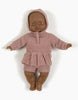 A Minikane Babies doll with medium brown skin is dressed in a Minikane Doll Clothing | Élie Tea Pink Knit Bonnet and matching shorts. The doll's eyes and lips are painted, showcasing the intricate details of the doll clothing, and it is positioned against a plain white background.