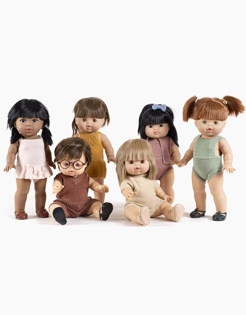 A group of six dolls with diverse appearances and outfits is arranged in two rows. The front row has three dolls sitting, and the back row has three standing. The dolls wear various colors of clothing, such as Minikane | Orlando Heather Caramel Knit Romper Doll Clothing, and some have accessories like glasses and hair bows.