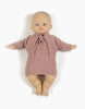 A Minikane Babies doll with a light skin tone is dressed in a hand-washable knitted pink outfit, featuring the Minikane Doll Clothing | Tea Pink Knit Alix Cardigan tied at the neck. The doll is laying flat on a white background.