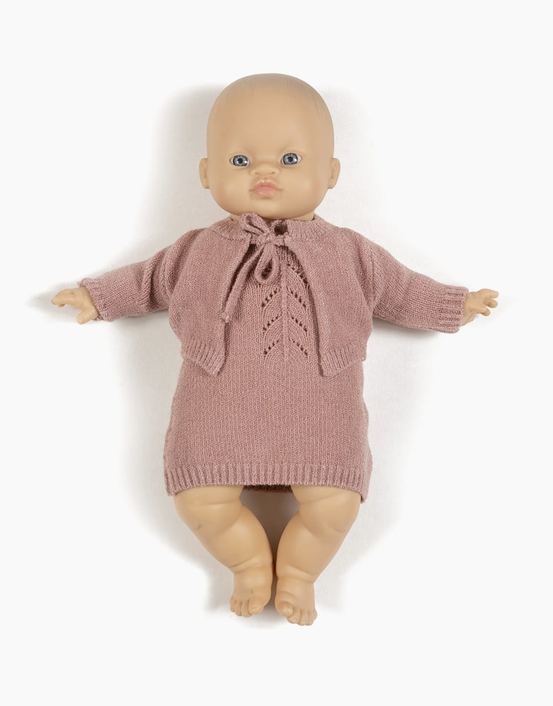 A Minikane Babies doll with a light skin tone is dressed in a hand-washable knitted pink outfit, featuring the Minikane Doll Clothing | Tea Pink Knit Alix Cardigan tied at the neck. The doll is laying flat on a white background.