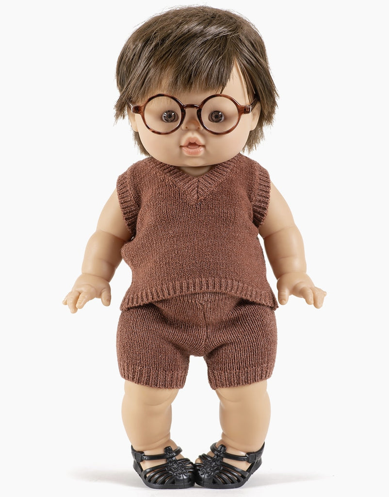 A Minikane Doll Clothing | Vito Shorts in Marl Caramel Knit with short brown hair, round glasses, and a neutral expression. The doll is dressed in a brown knit sleeveless top and matching knitted shorts made from a cozy wool composition, paired with black sandals. The background is plain and white.