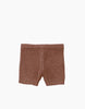 A pair of brown knitted shorts, featuring a subtle wool composition, laid flat against a white background. These Minikane Doll Clothing | Vito Shorts in Marl Caramel Knit have a simple design with an elastic waistband and a gentle knitted texture.